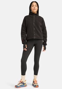 Giacca nera in pile con zip e colletto alto, abbinata a leggings neri e alla sneaker sportive multicolori con suole testurizzate.