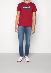 Červené bavlněné tričko s námořnickým logem Levi's, doplněné světle modrými denimovými džíny a bílými sneakers s červenými akcenty na jednoduchém pozadí.