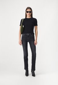 T-shirt noir ajusté, jean taille haute délavé foncé, sac à épaule vert, bottines noires. Le modèle se tient contre un fond blanc uni.