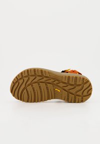 Suela de goma con un patrón de tracción grueso y texturizado en color marrón, que presenta un logo en amarillo. Diseñada para durabilidad y tracción.