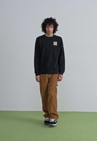 Zwarte sweatshirt met een klein beige label, gedragen met losse bruine broeken en zwarte sneakers. Eenvoudig ontwerp, relaxte pasvorm. Groene vloer.