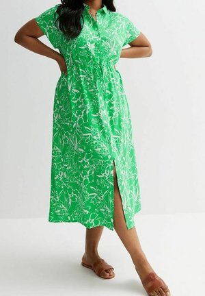 Kvinde iført en grøn blomsterprint midi-kjole med frontslids og brune flade sandaler, stående med hænderne på hofterne mod en hvid baggrund.