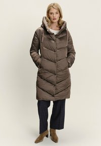 Smeđi kaput s podstavom dug do poda i kapuljačom, s uzorkom chevron i bočnim džepovima, nosi se preko tamnih kariranih hlača i čizama s visokom petom.