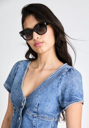 Occhiali da sole neri con montatura quadrata, dotati di lenti sfumate e dettagli metallici. Il modello indossa un vestito di jeans blu con maniche corte e scollo a V.
