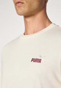 Beige bomull t-skjorte med rund hals, med rød brodert "PUMA" tekst og en grå puma-logo på venstre brystområde.