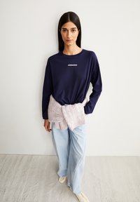 Marineblauwe longsleeve met witte tekst, gecombineerd met een lichtpaarse kabelgebreide trui om de taille geknoopt en lichtblauwe wijde jeans.