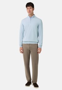 Pull-over bleu clair zippé en tissu doux, associé à un pantalon beige. Le pull présente des poignets et un ourlet côtelés, offrant une coupe décontractée.