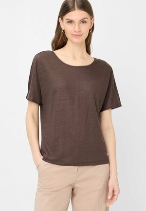 Vrouw met lang bruin haar, wearing gouden oorringen, een losse bruine korte mouw top en beige broek tegen een effen witte achtergrond.