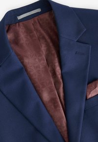 Dunkelblauer Blazer mit strukturiertem Äußeren, ausgestattet mit einer bordeauxfarbenen gemusterten Fütterung und einem passenden Einstecktuch. Verstellbare Ärmeldetails sind sichtbar.