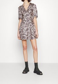 Vrouw in een korte bloemen wikkeljurk met pofmouwen en zwarte chunky enkellaarzen tegen een effen witte achtergrond.
