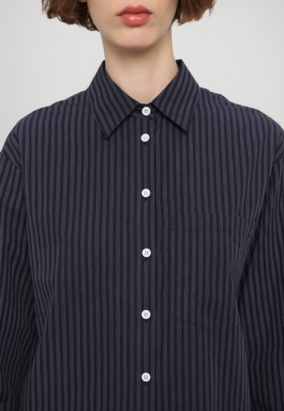 Camisa de rayas azul marino de botonadura en tela suave con botones blancos, un cuello y un bolsillo en el lado izquierdo. El patrón vertical añade textura.