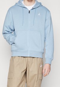 Ljusblå zip-hoodie i mjukt tyg med framfickor och justerbar dragsko på huvan; har en vit logotyp på bröstet.