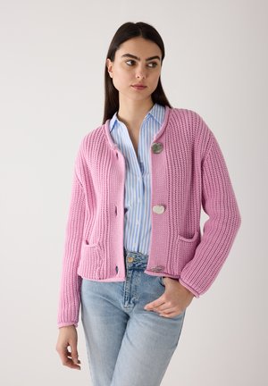 Femme portant un cardigan épais à gros boutons rose par-dessus une chemise rayée bleu et blanc et un jean bleu clair.