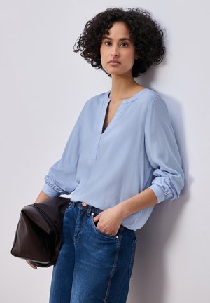 Femme aux cheveux bouclés portant un chemisier bleu clair et un jean, tenant un sac marron foncé, appuyée contre un mur blanc.