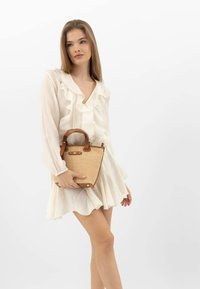 Spleciona torba ze słomy z brązowymi skórzanymi akcentami, prostokątny kształt, uchwyty do noszenia i szczegół logo w kolorze złotym. Nosi ją dziewczyna w białej sukience z falbanami.