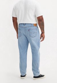 Man med ljusblå denimjeans, vit t-shirt, svarta sneakers med vita sulor, som står vänd bort från kameran på en helt vit bakgrund.