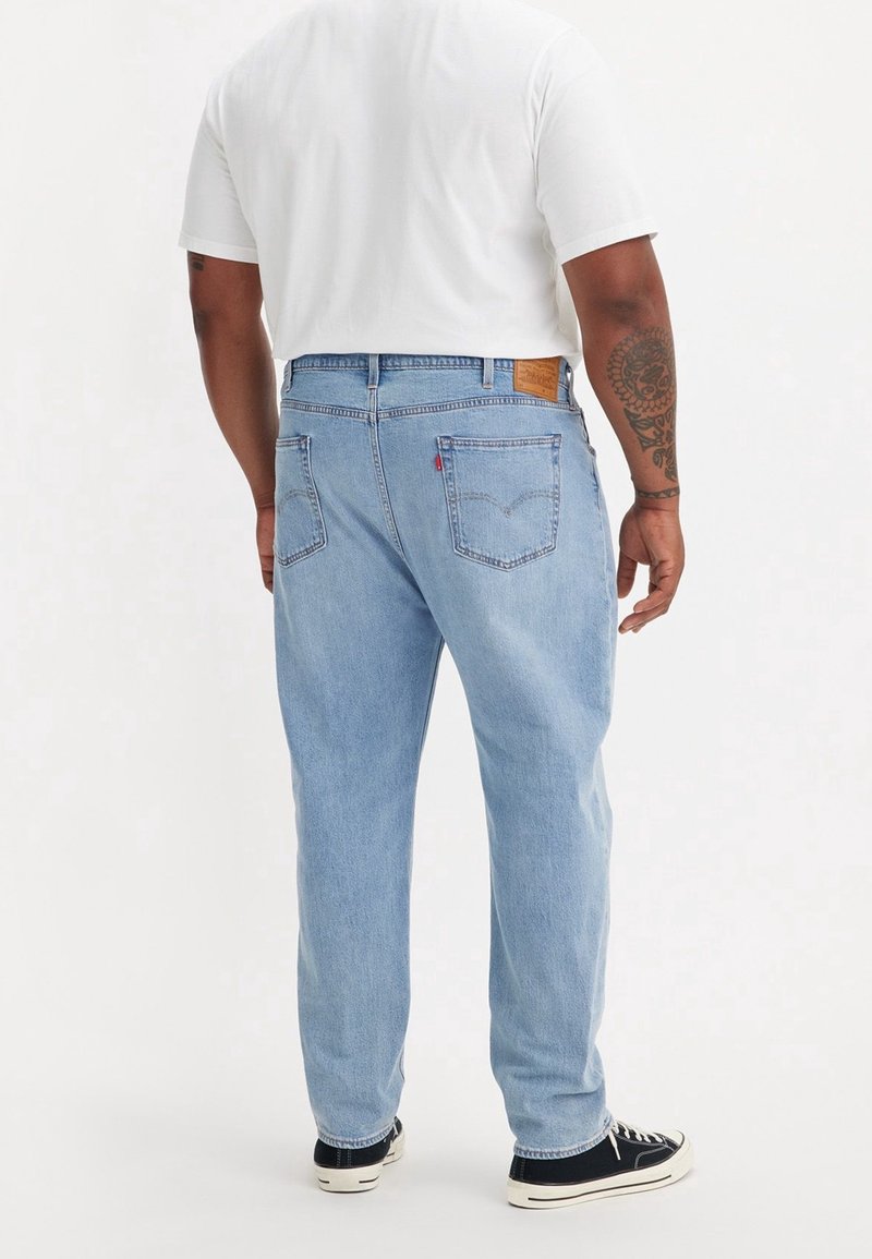 Man med ljusblå denimjeans, vit t-shirt, svarta sneakers med vita sulor, som står vänd bort från kameran på en helt vit bakgrund.