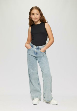 Jongedame met lang golvend haar, gekleed in een zwart mouwloos topje, lichtblauwe brede jeans en witte sneakers, staande met handen in haar zakken.
