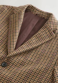 Pruunruuduline jakk tekstuurse kangaga, millel on tumepruun sisemine vooder ja ees üks must nööp.