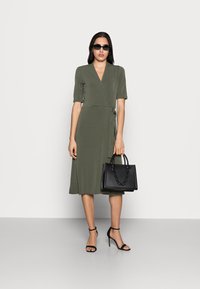 Vestido envolvente verde oliva com mangas curtas, um laço na cintura, combinado com uma mala preta texturizada e sapatos de salto alto pretos com tiras.