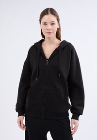 Sudadera negra con cremallera hecha de tela suave, que presenta una gran capucha, bolsillos frontales y puños acanalados. Textura suave con un diseño minimalista.