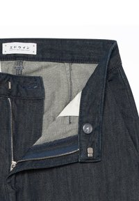 Jean en denim bleu foncé ouvert montrant la fermeture éclair, le bouton en métal, la fermeture à crochet et barre, et une étiquette intérieure blanche avec un texte japonais et l'URL du site web.