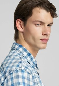 Camicia di jeans a quadri blu e bianco con collo e chiusura frontale a bottoni, indossata da un modello che guarda leggermente di lato.