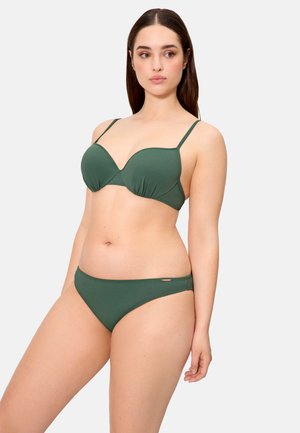 Mujer con bikini de dos piezas verde oscuro, de pie ligeramente girada, mirando de lado contra un fondo claro y liso.