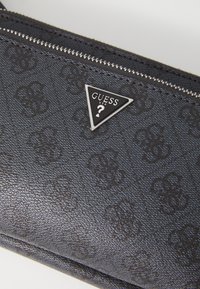 Portefeuille noir texturé avec un motif de logo répété, quincaillerie en métal argenté et une plaque triangulaire avec le détail "GUESS".