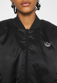 Svart bomberjacka med en slät, glänsande yta, med dragkedja framtill och rund hals. Har en vit broderad logga på vänster bröst.