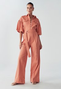 Mono de terracota con diseño de cuello y botones, mangas cortas abullonadas, cintura ceñida con cinturón y pantalones de pierna ancha. Textura suave.