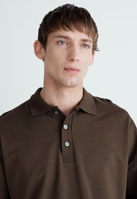 Brun polo skjorte laget av glatt stoff, med krage, tre-knapps placket og en liten logo på venstre side.