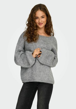 Giovane donna con capelli ricci che indossa un maglione grisaglia largo e jeans neri, sorridendo con le mani congiunte davanti a sé.