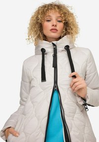 Veste matelassée gris clair avec un col haut, une fermeture éclair noire et des accents à cordon. Portée sur une chemise bleue à texture lisse.