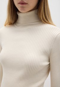 Maglione beige a collo alto con coste, caratterizzato da un design aderente e un colletto alto realizzato in un tessuto morbido e elastico, che mette in risalto una sottile texture.