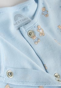 Body albastru deschis pentru bebeluși, realizat dintr-un material moale și texturat, cu ilustrații mici cu ursuleți și capse de fixare. Guler și mâneci cu margine reiată.