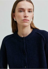 Marineblå cardigan med rund halslinje, knapper foran og strukturert ullstoff. Har raglanermer og svarte knapper for lukking.