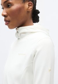 Ελαφριά μπεζ μπουφάν με κουκούλα, μαλακή υφή και διακριτική λεπτομέρεια λογότυπου στο στήθος, με ένα διακριτικό στοιχείο στο μανίκι.