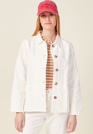 Witte denimjas met knoopsluiting, twee voorzakken en een casual pasvorm, gedragen over een multicolor gestreepte trui en witte broek.