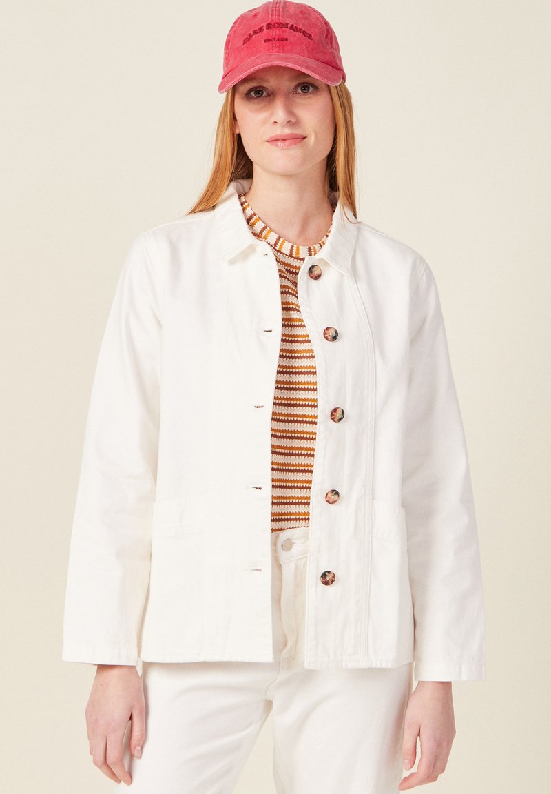 Giacca di denim bianco con chiusura a bottoni, due tasche frontali e vestibilità rilassata, indossata sopra un maglione a righe multicolori e pantaloni bianchi.