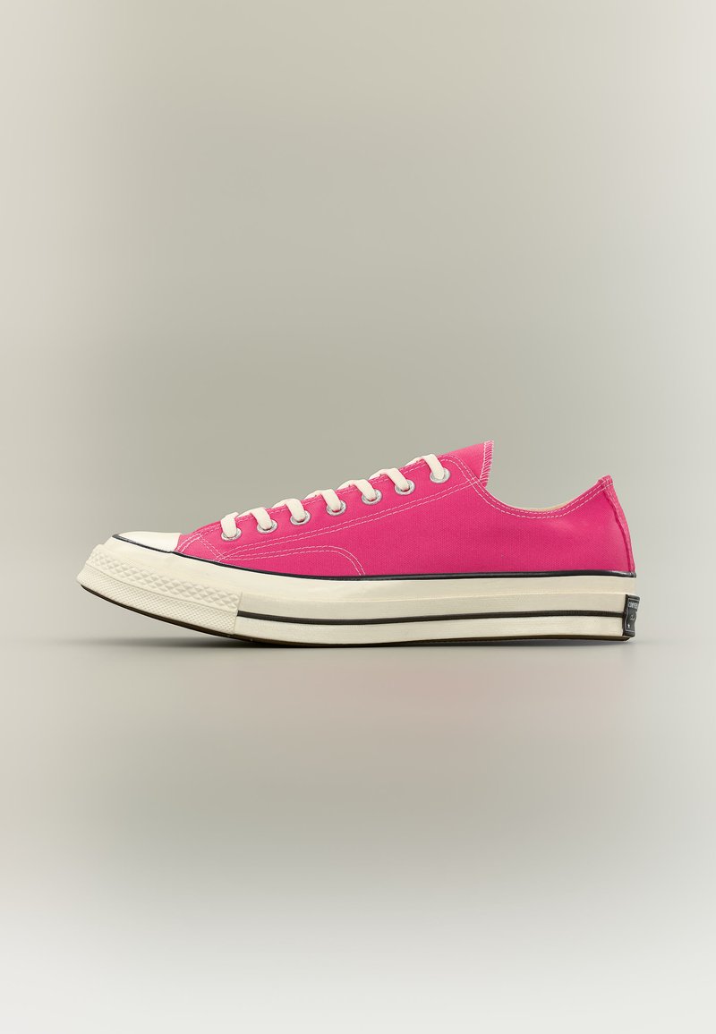 Niedriger pinker Leinensneaker mit weißer Gummisohle und Zehenkappe, weißen Schnürsenkeln und schwarzen Streifenakzenten auf neutralem Hintergrund.