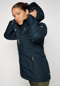 Veste isolée bleu marine avec une fermeture éclair à l'avant, capuche ajustable et poches latérales. Elle présente des accessoires marron et un petit détail de drapeau du pays.