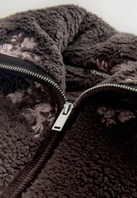 Ruda, pūkuota, tekstūrinė vilna su matomu sidabriniu užtrauktuku. Pasižymi rožinėmis gėlių siuvinėjimo detalėmis palei atidarymą.