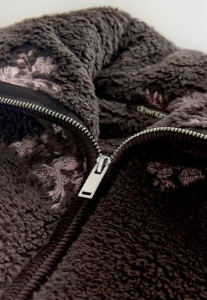 Brazs, pūkains, teksturēts vilnas audums ar redzamu sudraba rāvējslēdzēju. Pie ieejas ir rozā ziedu izšuvums.