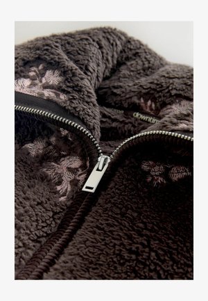 Brązowy, puszysty, teksturowany materiał polarowy z widocznym srebrnym zamkiem błyskawicznym. Posiada różowe hafty kwiatowe wzdłuż otwarcia.