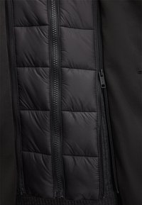 Giacca nera con zip frontale e imbottitura trapuntata. Materiale esterno liscio con una morbida texture interna e dettagli con doppia zip.