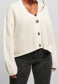 Femme portant un cardigan blanc épais boutonné sur un top noir et un jean noir taille haute, avec des mèches de cheveux bouclés visibles.