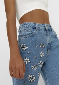 Blå denimjeans med broderade vita prästkragar på framsidan, med hög midja och avslappnad passform. Vit croptop delvis synlig.