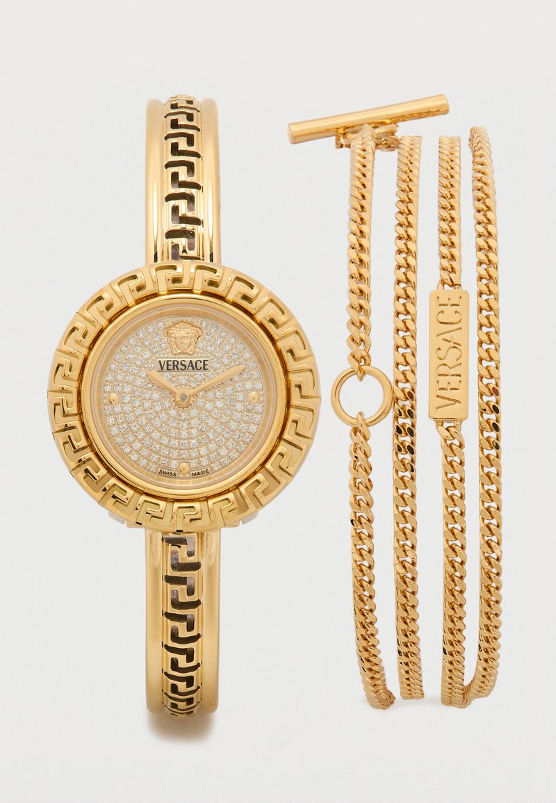 Relógio Versace com pulseira de metal dourado, mostrador redondo, adornado com cristais transparentes, apresentando o logótipo da marca e detalhes geométricos. Inclui três correntes douradas.