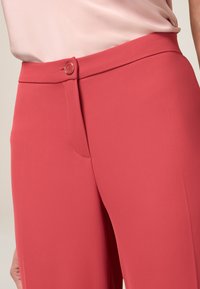 Dopasowane spodnie w intensywnym koralowym kolorze, wykonane z gładkiego materiału. Posiadają zapięcie na guzik, ukrytą przednią kieszeń oraz dopasowany krój.